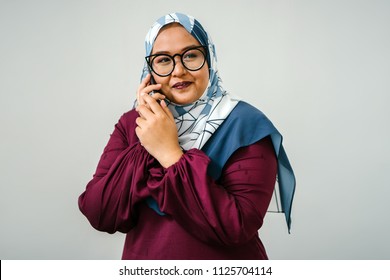 Portrait Of A Muslim Malay Woman Talking On A Smartphone. She Is Against A Plain White Background (studio) And Is Fashionably Dressed In A Colorful Tudung Head Scarf And Traditional Orange Garb. 