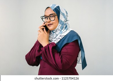 Portrait Of A Muslim Malay Woman Talking On A Smartphone. She Is Against A Plain White Background (studio) And Is Fashionably Dressed In A Colorful Tudung Head Scarf And Traditional Orange Garb. 