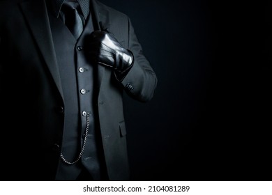 Portrait Of Mysterious Man In Dark Suit And Leather Gloves Standing Proudly On Black Background. Concept Of Mafia Hitman Or Gentleman Assassin.