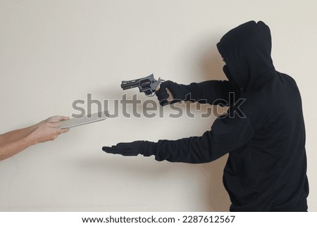 Portrait of mysterious man wearing black hoodie and mask extorting a laptop from victim. Isolated image on gray background
