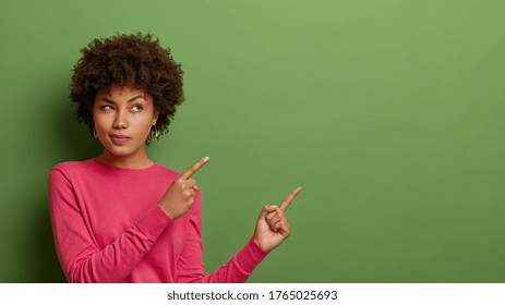 Portrait of mysterious thoughtful woman with curly Afro hair, points away on blank space, dressed in casual pink jumper, isolated on green background. You should see this. People and advertisement - Shutterstock ID 1765025693