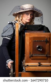 Portrait of a mysterious young positive woman with a modest long dress and hat posing near an old daguerreotype camera. Concept stylization of retro vintage photo.