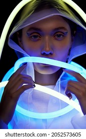 Portrait Of Mystic Woman In White Transparent Raincoat And Flexible Blue And White Neon Tube Lights. Black Background                                                      