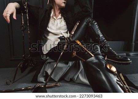 Portrait of a Mystical or magical girl with a gothic make-up and auburn hair, dressed in a long coat, off-white shirt, tight leather leggings and tall laced chunky boots. Posing in a dark gothic room.