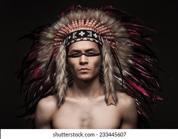 Portrait Of The Naked Indian Strong Man Posing With Traditional Native American Make Up  And Headdress Looking At The Camera. Close Up Desaturated Studio Shot