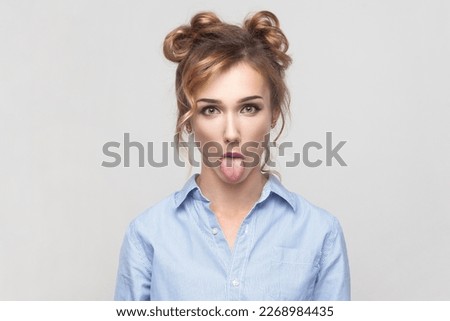 Portrait of naughty comic blonde woman sticking out tongue, misbehaves, demonstrating her disobedience and protest, has funny look, wearing blue shirt. Indoor studio shot isolated on gray background.