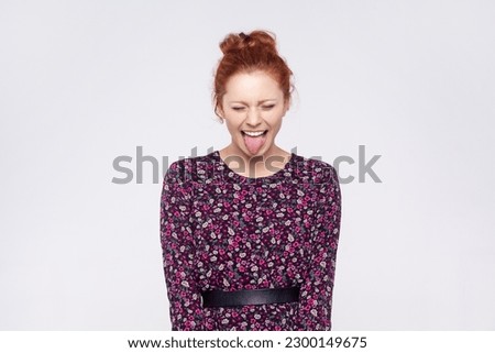 Portrait of naughty playful ginger woman with bun hairstyle wearing dress showing tongue at camera with closed eyes, having fun, joke. Indoor studio shot isolated on gray background.