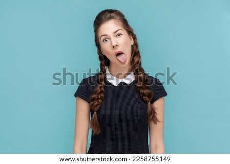 Portrait of naughty woman with braids sticking out tongue, making funny grimace, looking and winking at camera, wearing black dress. woman Indoor studio shot isolated on blue background.