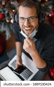 Portrait Of Nerd Man With Christmas Tree On The Background Holding Phone Smartphone Above The Laptop During The Covid-19 Pandemic With Face Mask At Home Office