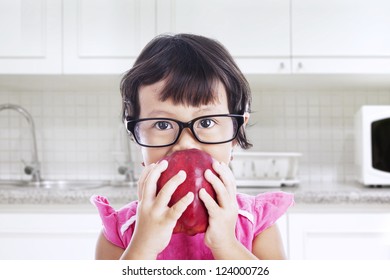 Portrait Of Nerd Toddler Is Eating Red Apple In The Kitchen