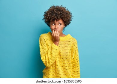 Portrait of nervous Afro American woman keeps hands near mouth feels concerned before important interview hesitates about something dressed in knitted yellow jumper poses against blue studio wall