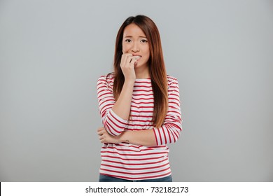 Portrait of a nervous asian girl biting her nails while looking at camera isolated over gray background