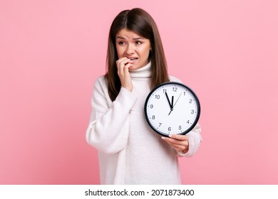 Portrait of nervous brunette young female holding big wall clock, biting fingernails, looking away, wearing white casual style sweater. Indoor studio shot isolated on pink background.