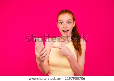 Portrait of nice cheerful positive dreamy eager red-haired girl in tanktop with ponytail, getting congrats sms, poining at smart-phone, isolated on bright vivid fuchsia background