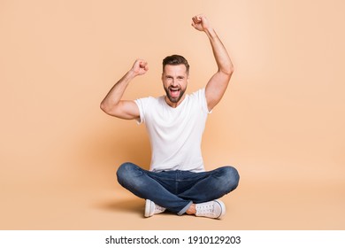 Portrait of nice cheery guy sitting on floor rejoicing victory triumph isolated over beige color background