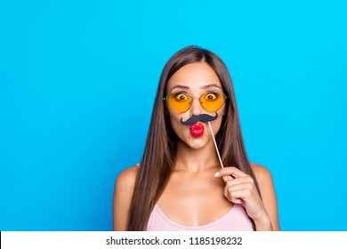 Portrait of nice cool winsome adorable positive optimistic straight-haired girl wearing tanktop and yellow sun glasses, sending air kiss, trying fake mustache, isolated on bright vivid blue background