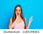 Portrait of nice positive optimistic attractive adorable stylish straight-haired girl wearing yellow sunglasses, tanktop, pointing aside, isolated over bright vivid blue background, red lips pomade