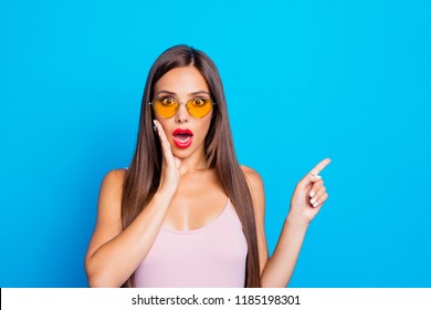 Portrait of nice positive optimistic attractive adorable stylish straight-haired girl wearing yellow sunglasses, tanktop, pointing aside, isolated over bright vivid blue background, red lips pomade