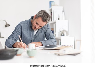 Portrait Of A Nice Smiling Grey Hair Man With Beard, Working At Home On Some Project, He Is Sitting At A White Table, Writing Creative Ideas On Papers With Tea In Front Of Him. Focus On The Man