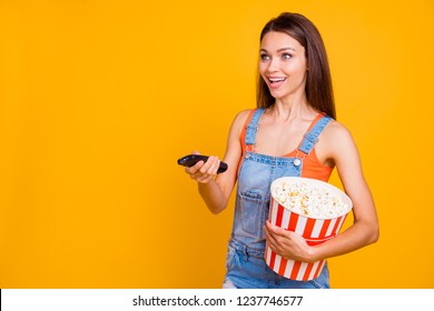 Portrait of nice sweet lovely adorable charming attractive cheerful glad girl wearing overall holding in hands popcorn box pressing button on controler isolated on bright vivid shine yellow background