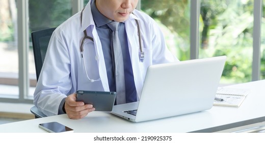 Portrait Of No Face Asian Senior Doctor Sitting In Medical Office While Using Laptop Computer And Tablet. Copy Space. Ratio 2:1