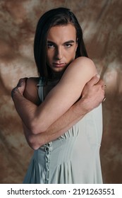 Portrait Of Nonbinary Person In Sundress Touching Shoulders On Abstract Brown Background