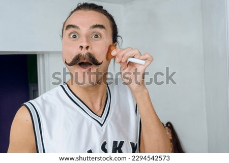 portrait of non-binary young latino person with mustache and collected hair, he is in his room looking at the camera with a funny face while doing makeup with a brush, lifestyle concept, copy space