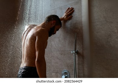 Portrait Of Nude Guy Taking Shower Standing Under Steaming Hot Water Washing Body And Relaxing In Modern Bathroom At Home, Leaning On Wall. Male Beauty, Bodycare And Everyday Hygiene For Men Concept