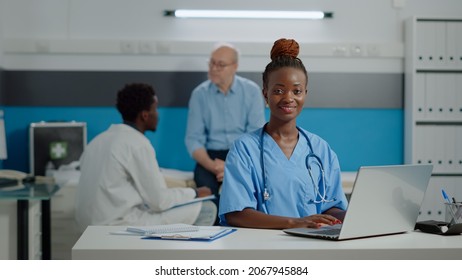 Portrait Of Nurse Typing On Laptop Keyboard In Medical Office At Facility. Assistant Woman With Uniform Sitting At Desk Using Gadget With Technology. Doctor And Old Patient In Background