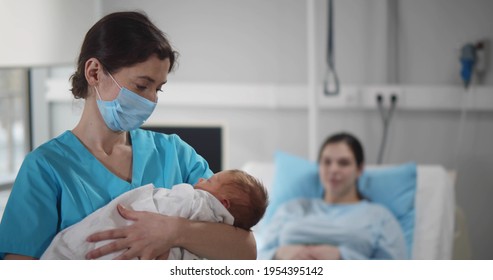 Portrait of nurse wearing safety mask cradling newborn in arms with young mother resting in bed at hospital ward. Happy woman relaxing in bed after childbirth with doctor holding baby