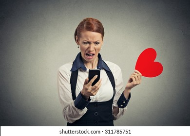 Portrait offended displeased young woman reading news on smart phone throwing away red heart isolated on gray wall background. Human facial expression emotion feeling reaction