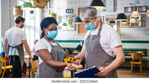 Portrait of old Caucasian man worker in cafeteria measuring temperature of young African American beautiful woman colleague in medical mask using infrared thermometer writing symptoms, health check - Powered by Shutterstock