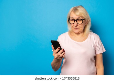 Portrait Of An Old Friendly Woman In Glasses And A Casual T-shirt Holding A Phone In Her Hands And Looking At The Screen On An Isolated Blue Background. Emotional Face