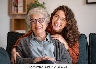 Portrait of old grandma and adult granddaughter hugging with love on sofa while looking at camera. Happy young woman with eyeglasses hugging from behind older grandma with spectacles generation family - Shutterstock ID 1910306023