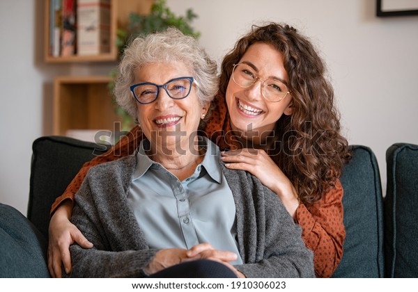 Portrait of old grandma and adult granddaughter\
hugging with love on sofa while looking at camera. Happy young\
woman with eyeglasses hugging from behind older grandma with\
spectacles generation\
family