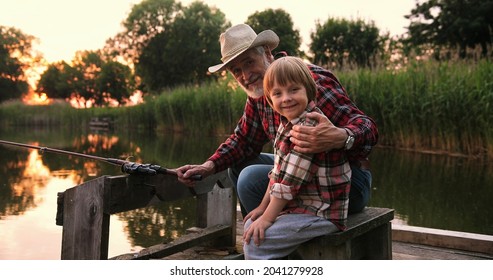 Portrait of the old gray haired fisherman with his grandson sitting at the lake at the pier, looking at each other and then smiling to the camera. Family relationships concept 