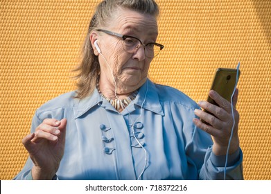 portrait of old lady, grandmother in shock. outdoors at sun day. modern caucasian granny in glasses listening music in headphone, earphones, headphones from phone. she has smartphone in her adult hand