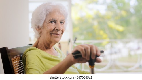 Portrait Of Old Lady Sitting On Rocker At Home. Happy Elderly Woman Relaxing On Rocking Chair And Holding Walking Stick. Copy Space