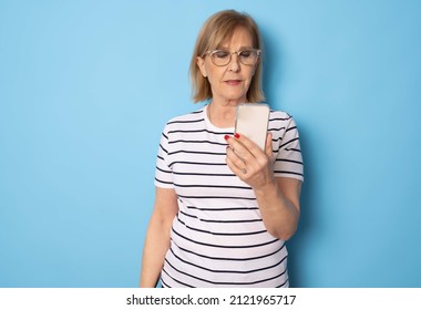 Portrait Of Old Lady Wearing Striped T-shirt Using Mobile Phone Standing Isolated Over Blue Background. Technology Concept.