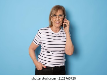 Portrait Of Old Lady Wearing Striped T-shirt Using Mobile Phone Standing Isolated Over Blue Background. Technology Concept.