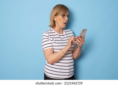 Portrait Of Old Lady Wearing Striped T-shirt Using Mobile Phone Standing Isolated Over Blue Background. Technology Concept.