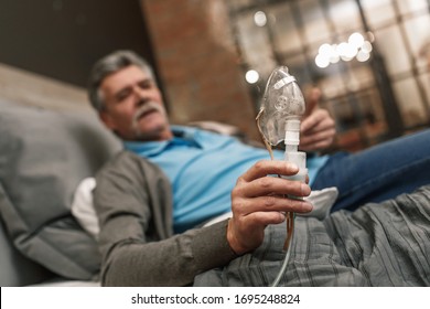 Portrait of old male holding inhalation through oxygen mask at home bedroom and use his laptop.