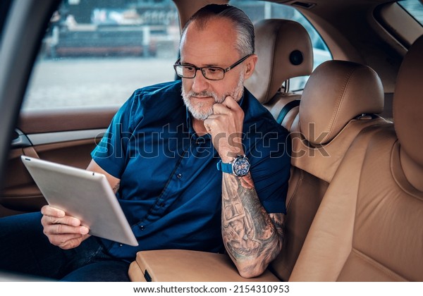 Portrait of old man with tattoo and glasses\
using tablet sitting inside of car in\
city.