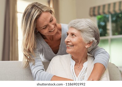 Portrait of old mother and mature daughter hugging at home. Happy senior mother and adult daughter embracing with love on sofa. Cheerful woman hugging from behind older mom and looking at each other.