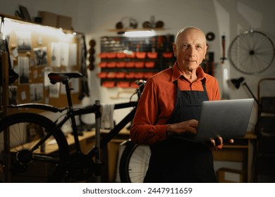 Portrait of a old repairman in workwear holding laptop computer at the bicycle workshop. Copy space - Powered by Shutterstock