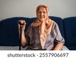 Portrait of an old sad Muslim woman with her walking stick at home wearing traditional hijab, looking at the camera.