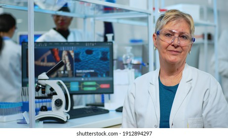 Portrait Of Old Scientist Woman Smiling At Camera In Modern Equipped Lab. Multiethnic Team Examining Virus Evolution Using High Tech And Chemistry Tools For Scientific Research, Vaccine Development.