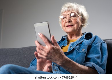 Portrait of old smiling lady looking at screen of mobile phone while sitting on couch