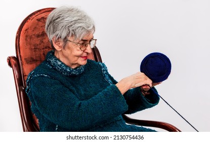 Portrait Of An Old Woman Sitting On A Rocker And Making A Ball Of Thread Against A White Background.