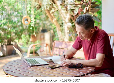 Portrait Of Older Asian Man In Red T Shirt Sitting On The Wooden Chair Writing A Diary Book With Computer, Smart Phone And Eyeglasses On The Table, In The Garden